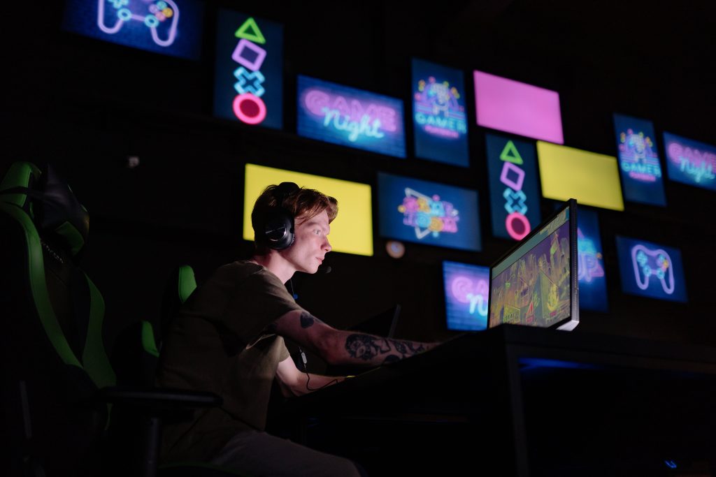 Gamer with headset playing video games on computer monitor surrounded by colorful gaming displays and PlayStation controll...